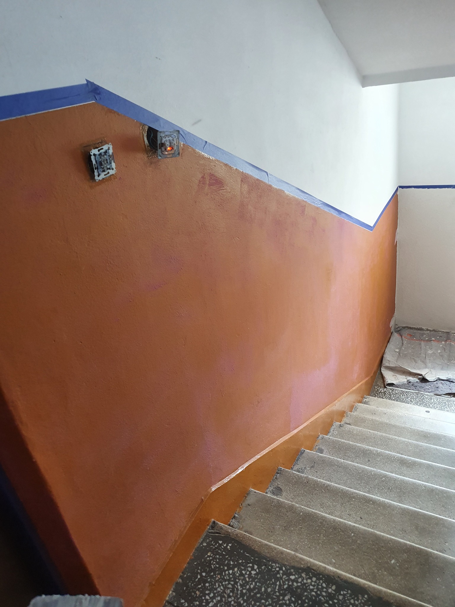 Malowanie klatki schodowej w Lubaczowie, widoczne zabezpieczone taśmą malarską krawędzie, odsłonięte puszki instalacyjne i świeżo pomalowana ściana w odcieniach brązu.