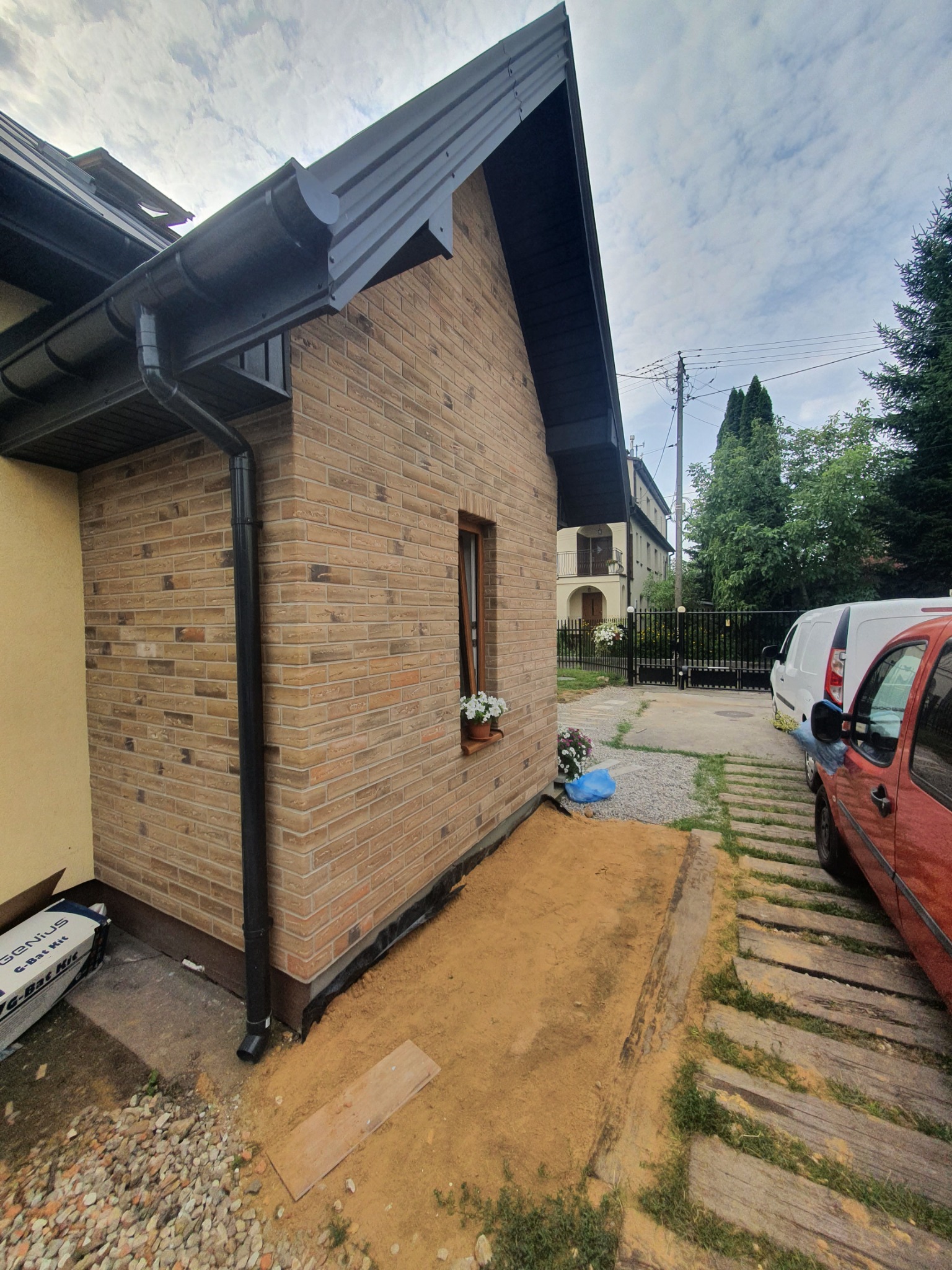 Fragment domu z elewacją z cegły klinkierowej, widoczny system rynnowy w kolorze antracytowym, okno z kwiatami w doniczce, teren wokół domu przygotowany pod prace ziemne.