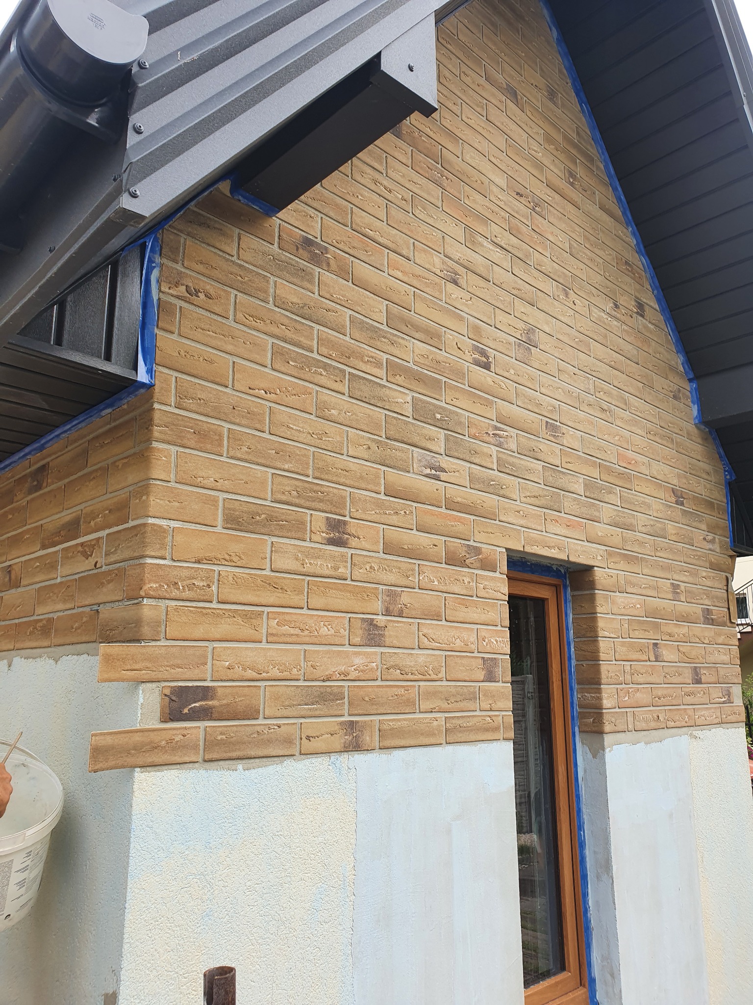 Kładzenie ceglanych płytek elewacyjnych na ścianie budynku, zabezpieczonej niebieską taśmą malarską wokół okna i pod dachem; widoczny fragment dłoni z wiadrem kleju.