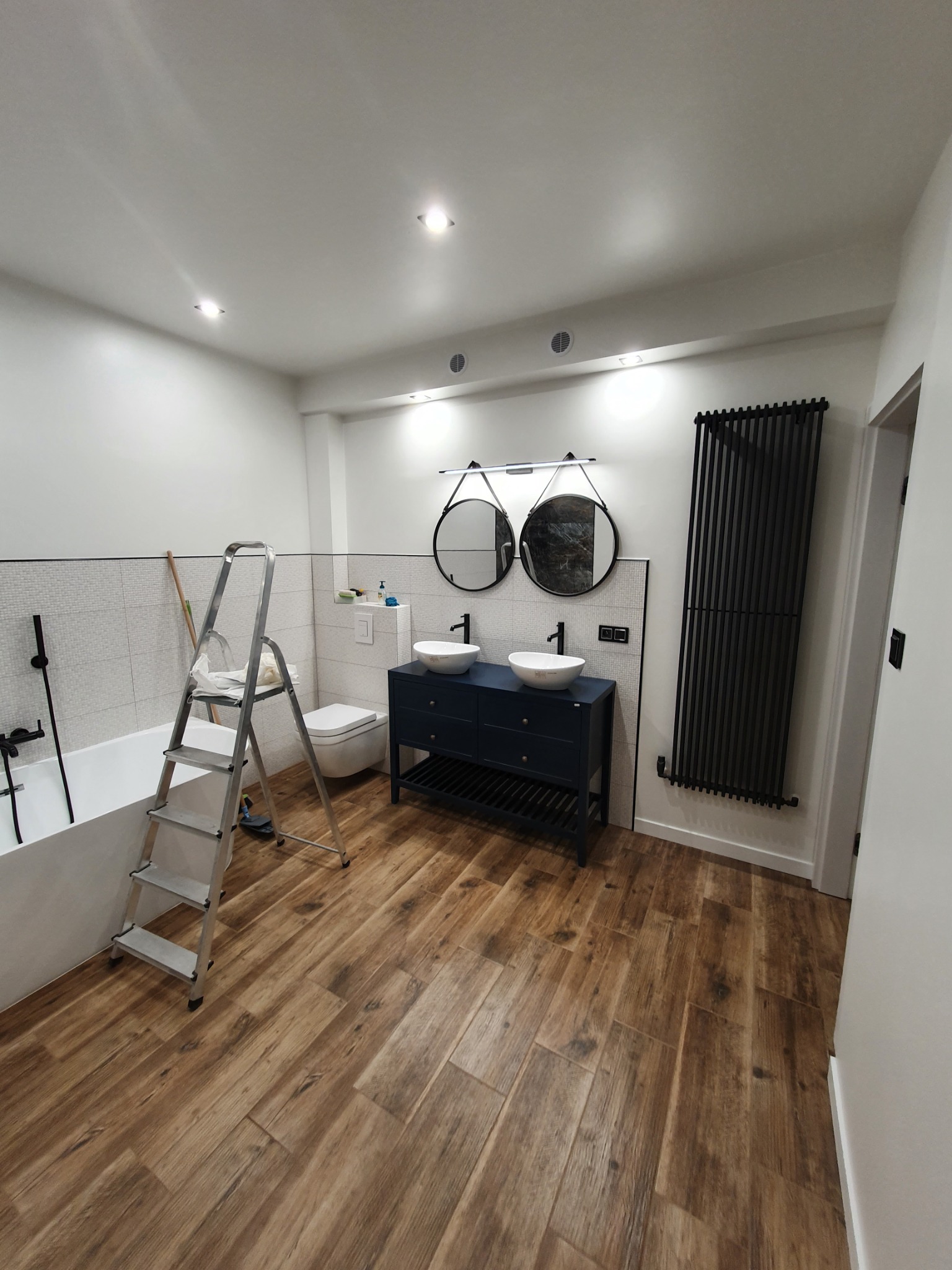 Nowoczesna łazienka z drewnopodobną podłogą, podwójną umywalką nablatową na granatowej szafce, czarnym grzejnikiem drabinkowym i drabiną opartą o wannę przy ścianie z mozaiką.