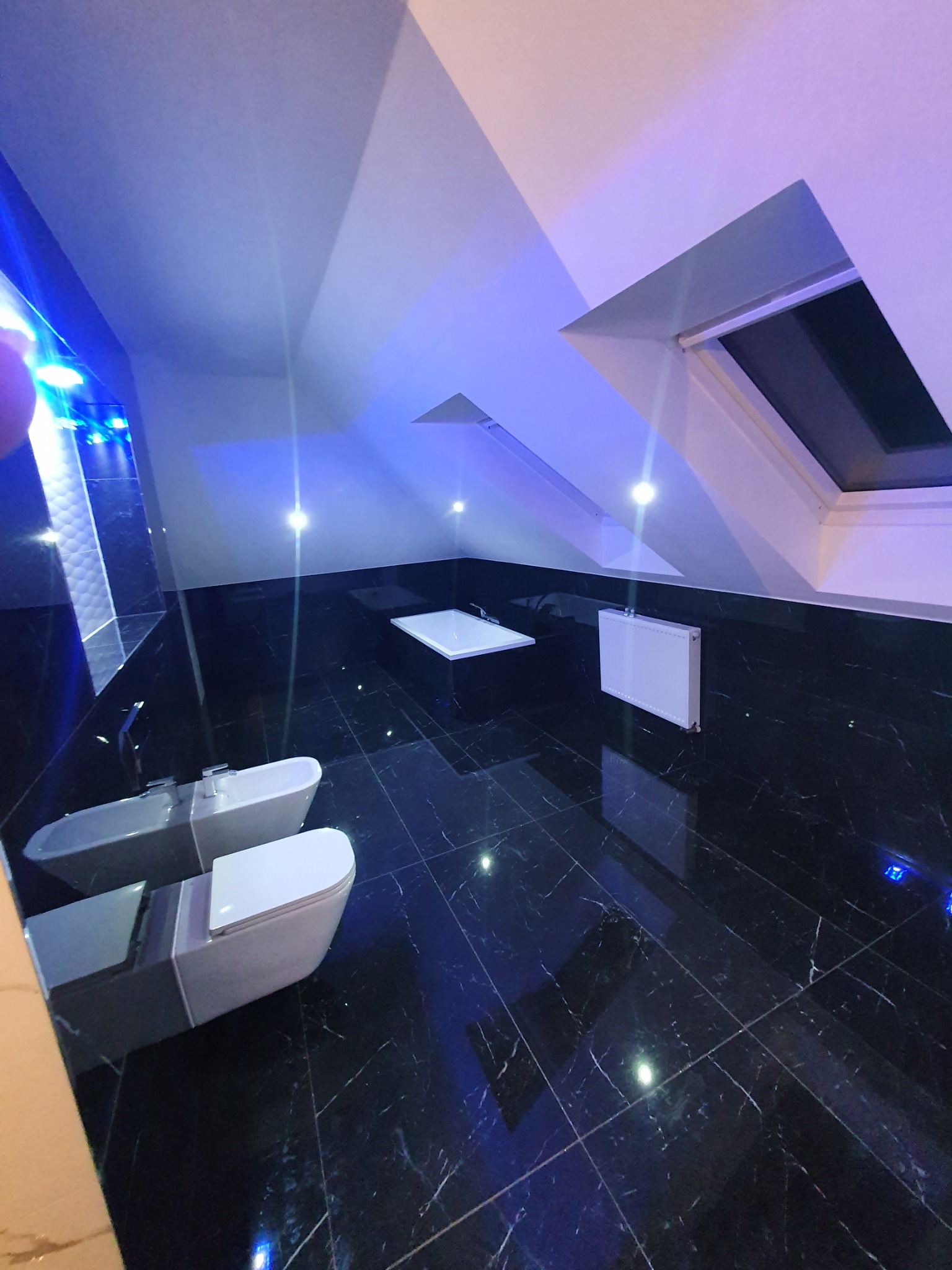 Nowoczesna łazienka na poddaszu z czarną marmurową podłogą, białą wanną, umywalką i toaletą, oświetlona niebieskim światłem LED.