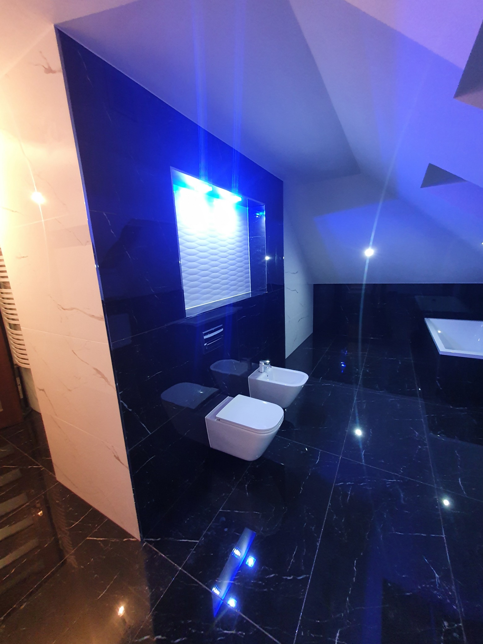 Nowoczesna łazienka na poddaszu z czarnymi marmurowymi płytkami, białymi sanitariatami i podświetleniem LED.