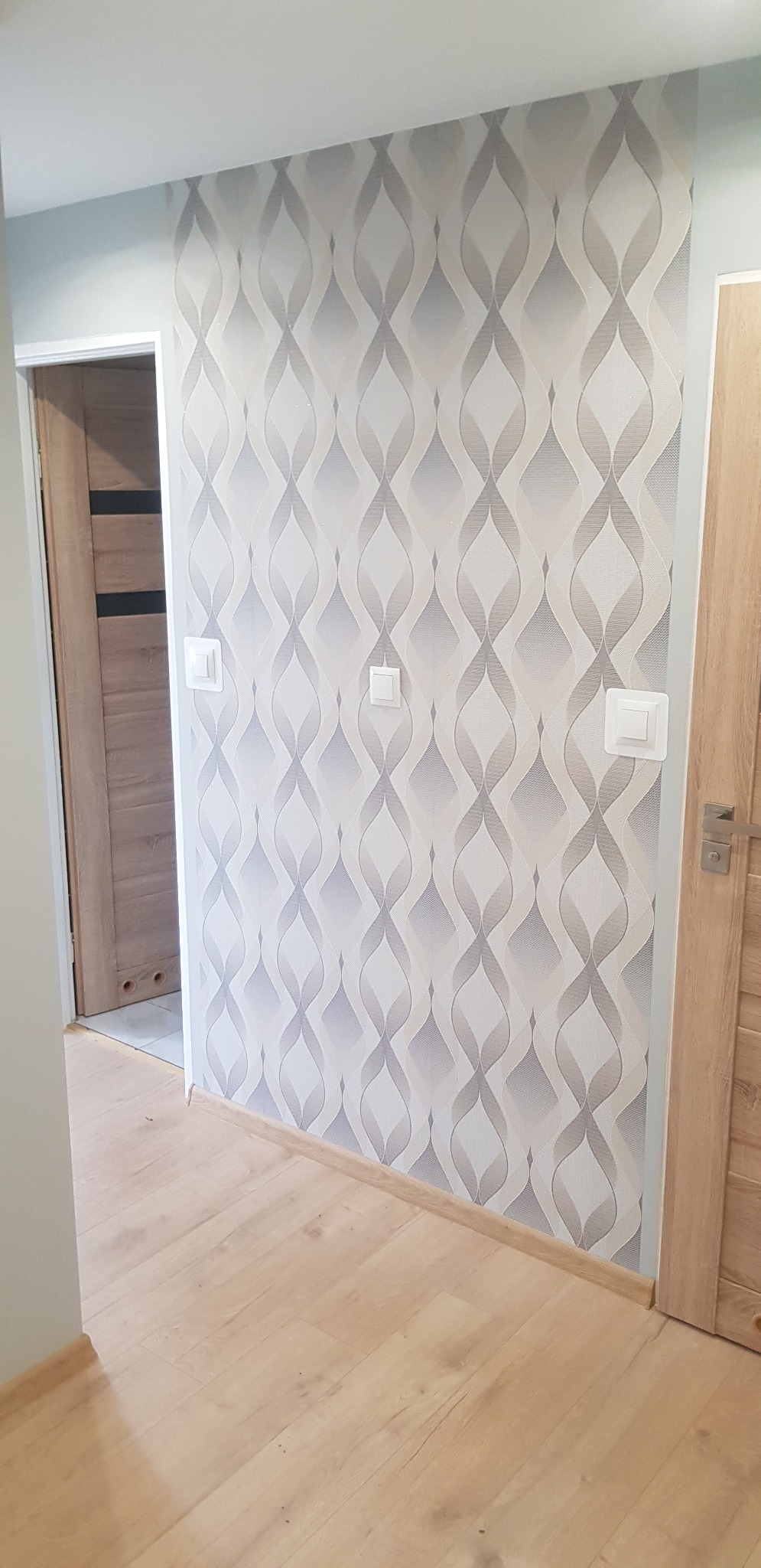 Wnętrze korytarza po remoncie: ściana pokryta tapetą w szaro-białe wzory, jasna drewniana podłoga, drzwi po lewej stronie z ciemniejszymi wstawkami, po prawej stronie jasne drzwi z metalową klamką,...
