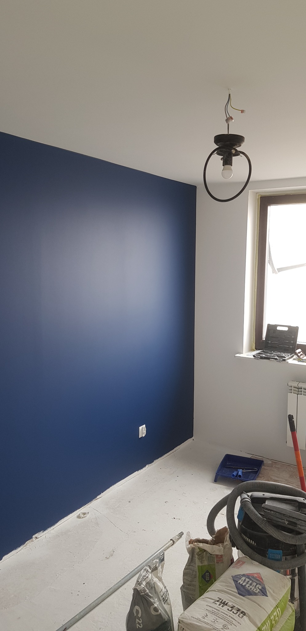 Świeżo pomalowany pokój z jedną ścianą w kolorze granatowym, widoczny sufit, podłoga przygotowana do dalszych prac wykończeniowych, widoczne worki z materiałami budowlanymi i odkurzacz przemysłowy,...