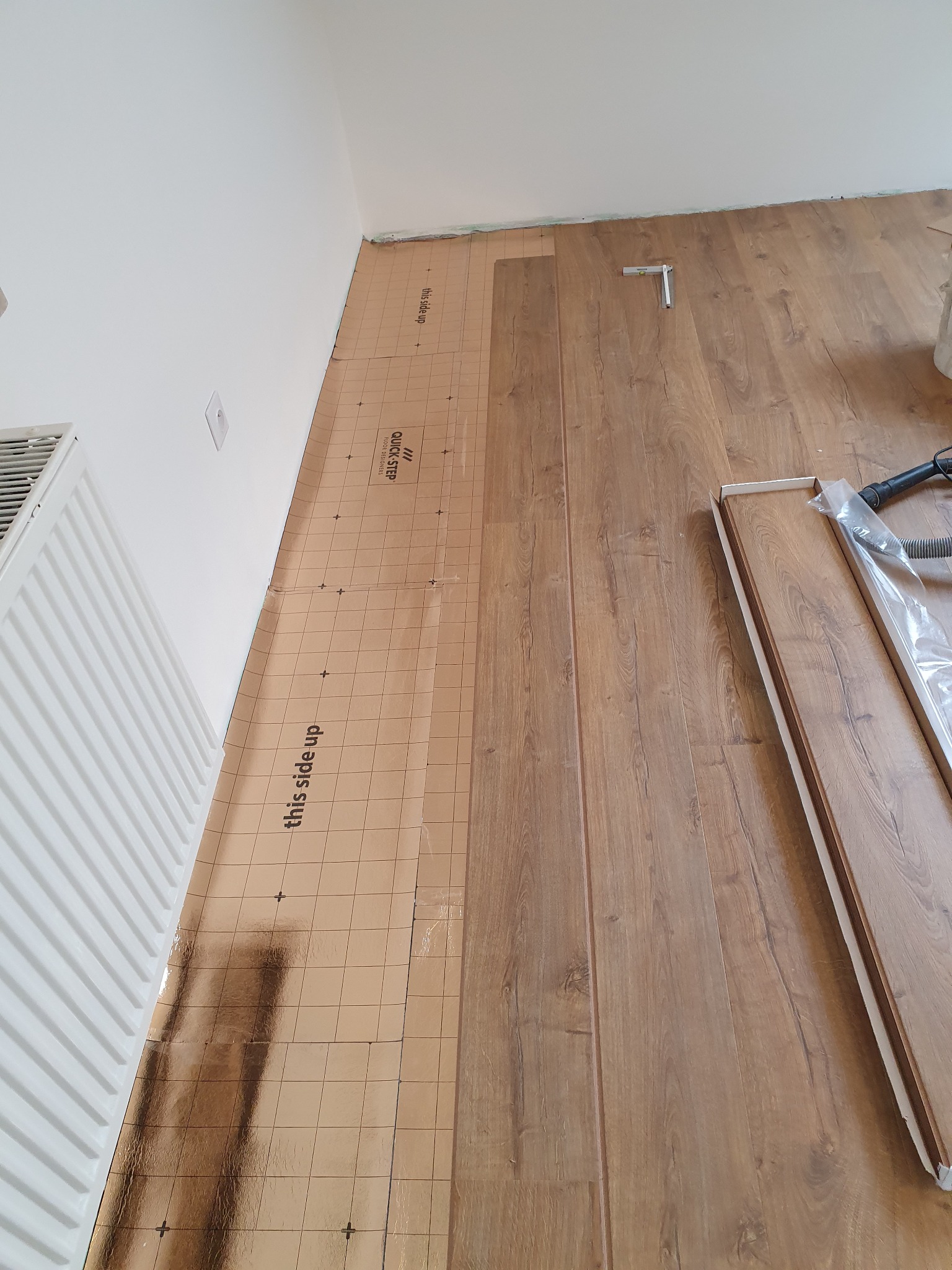 Montaż paneli podłogowych: widoczny podkład z nadrukiem 'this side up', częściowo ułożone panele, poziomica i odkurzacz w rogu pomieszczenia.