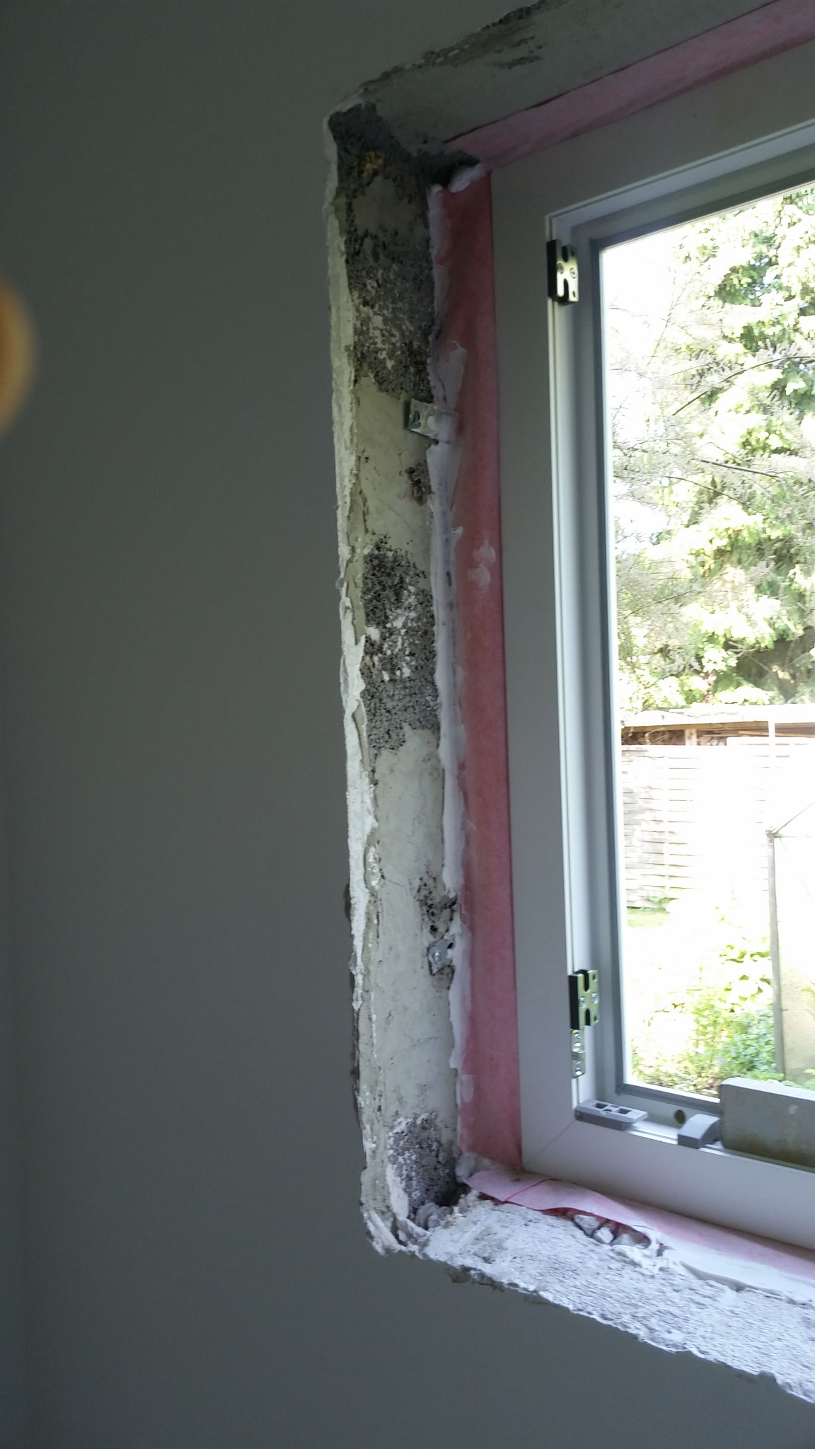 Przygotowanie ościeżnicy okiennej do dalszych prac wykończeniowych: surowy beton, resztki starej zaprawy i różowa folia izolacyjna widoczne wokół nowego, białego okna PCV.