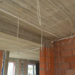 Surowy betonowy strop z widocznymi przewodami elektrycznymi zamocowanymi do sufitu i ścian w nowo budowanym domu z czerwonej cegły, wisząca żarówka na kablu.