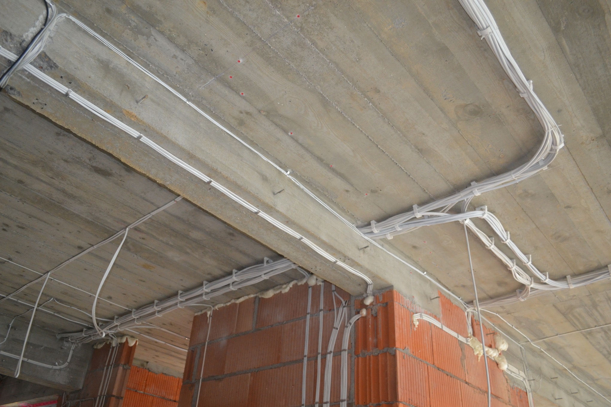 Białe kable elektryczne prowadzone po betonowym suficie i ceglanej ścianie w surowym stanie, mocowane za pomocą plastikowych uchwytów i pianki montażowej.