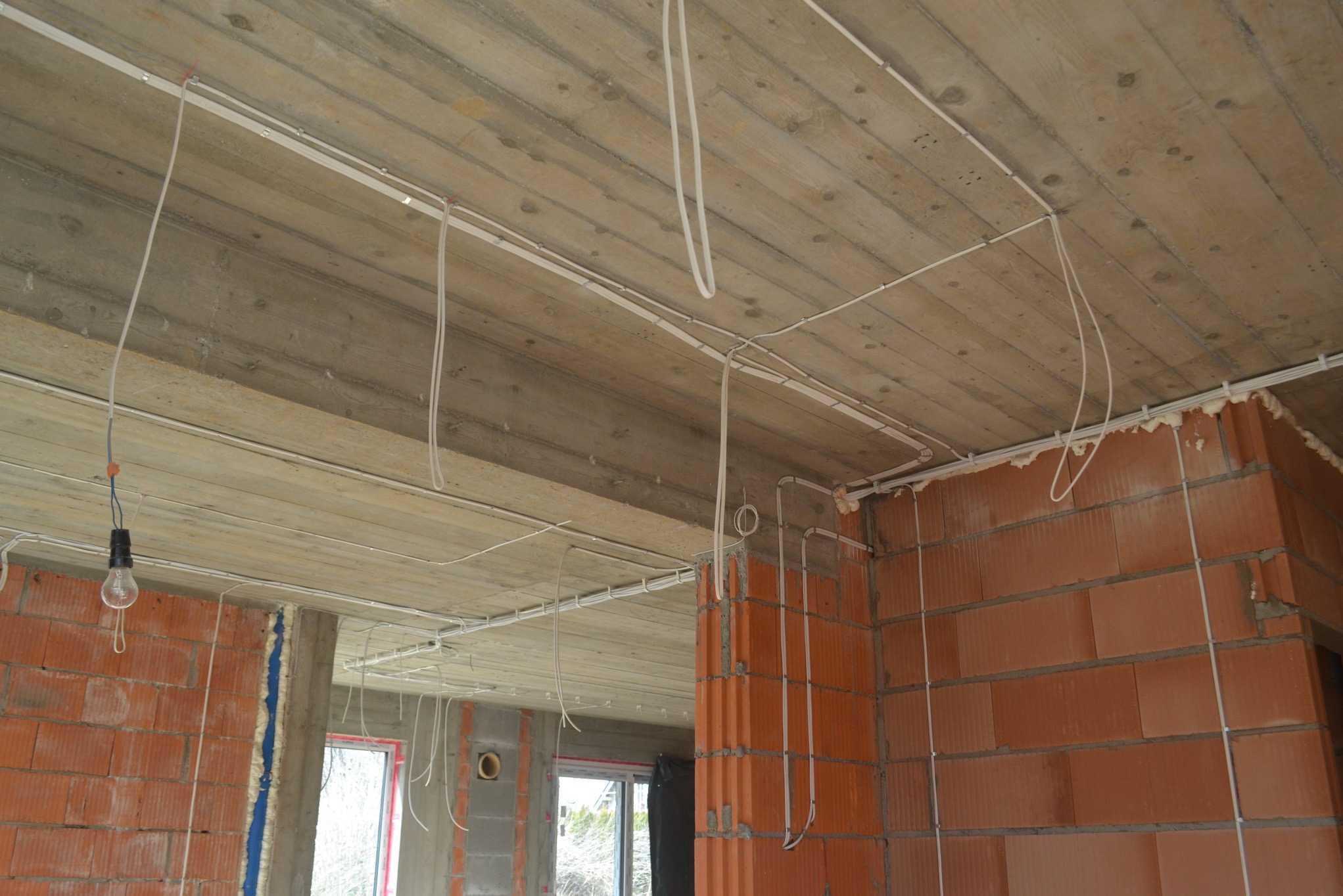 Surowy betonowy strop z widocznymi przewodami elektrycznymi zamocowanymi do sufitu i ścian w nowo budowanym domu z czerwonej cegły, wisząca żarówka na kablu.
