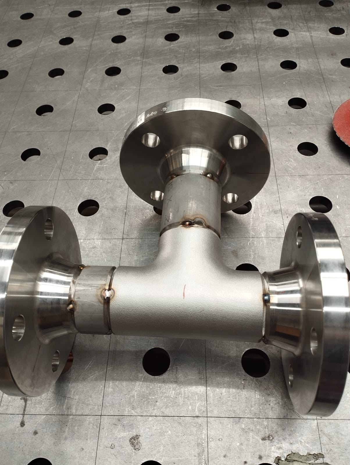 Metalowa rura w kształcie litery T z trzema kołnierzami połączonymi spawami, leżąca na metalowym stole spawalniczym z otworami.