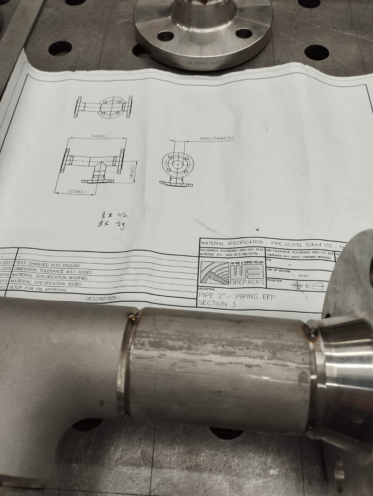 Zespawany element rurociągu ze stali nierdzewnej, widoczne spawy, leżący na stole warsztatowym obok rysunku technicznego z wymiarami i specyfikacją materiałową.
