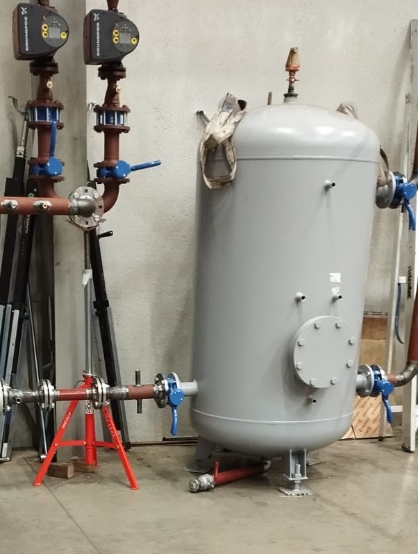 Szary zbiornik ciśnieniowy z armaturą, rurociągiem i pompami Grundfos w pomieszczeniu technicznym, wsparty na czerwonym stojaku montażowym.