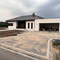 P.P.H.U BUD-MAX BOBULA JÓZEF - Nowoczesny dom jednorodzinny z utwardzonym podjazdem z kostki brukowej, widoczne obrzeża, w tle budynek w surowym stanie, pochmurne niebo.