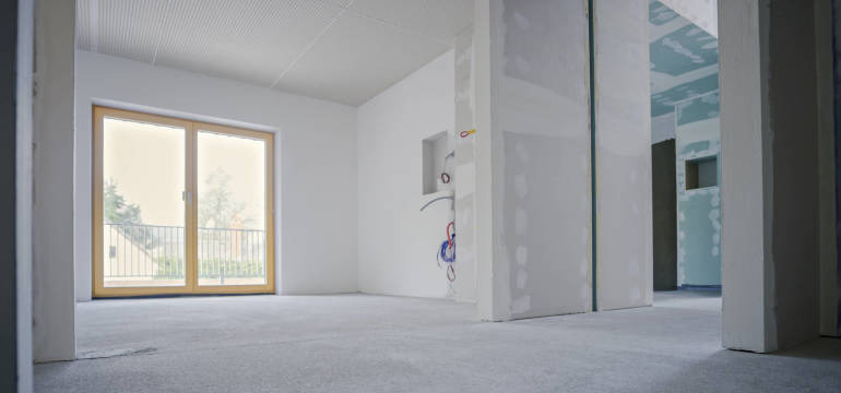 Wnętrze surowego stanu z drewnianą stolarką okienną, widoczne przewody elektryczne wystające ze ściany i ściany przygotowane pod malowanie.