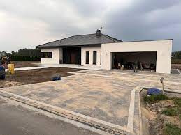 Nowoczesny dom jednorodzinny z utwardzonym podjazdem z kostki brukowej, widoczne obrzeża, w tle budynek w surowym stanie, pochmurne niebo.