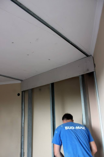 Pracownik w niebieskiej koszulce BUD-MAX montuje płytę gipsowo-kartonową na metalowym stelażu przy suficie w rogu pomieszczenia.