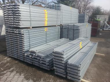 Stosy ocynkowanych, metalowych podestów rusztowaniowych z żółtymi paskami, ułożone na zewnątrz na kostce brukowej, w tle drzewa.