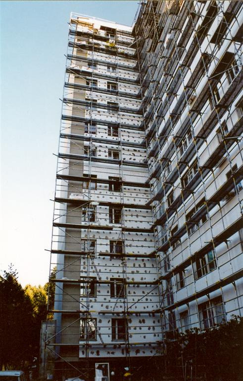 Wielopiętrowy budynek w trakcie ocieplania, osłonięty rusztowaniem, widoczne płyty izolacyjne mocowane do elewacji, ujęcie z dołu.