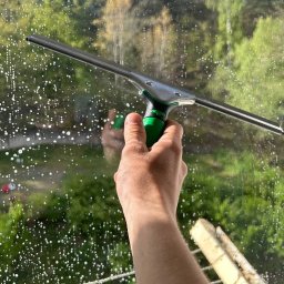 CleanTime - Ręka z zielonym uchwytem ściągaczki do okien na tle szyby pokrytej pianą, z widokiem na zieleń drzew i fragment drabiny sznurkowej w tle.