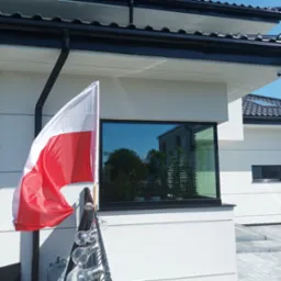 Biała elewacja domu z czarną stolarką okienną, na pierwszym planie flaga Polski i girlanda z żarówek.
