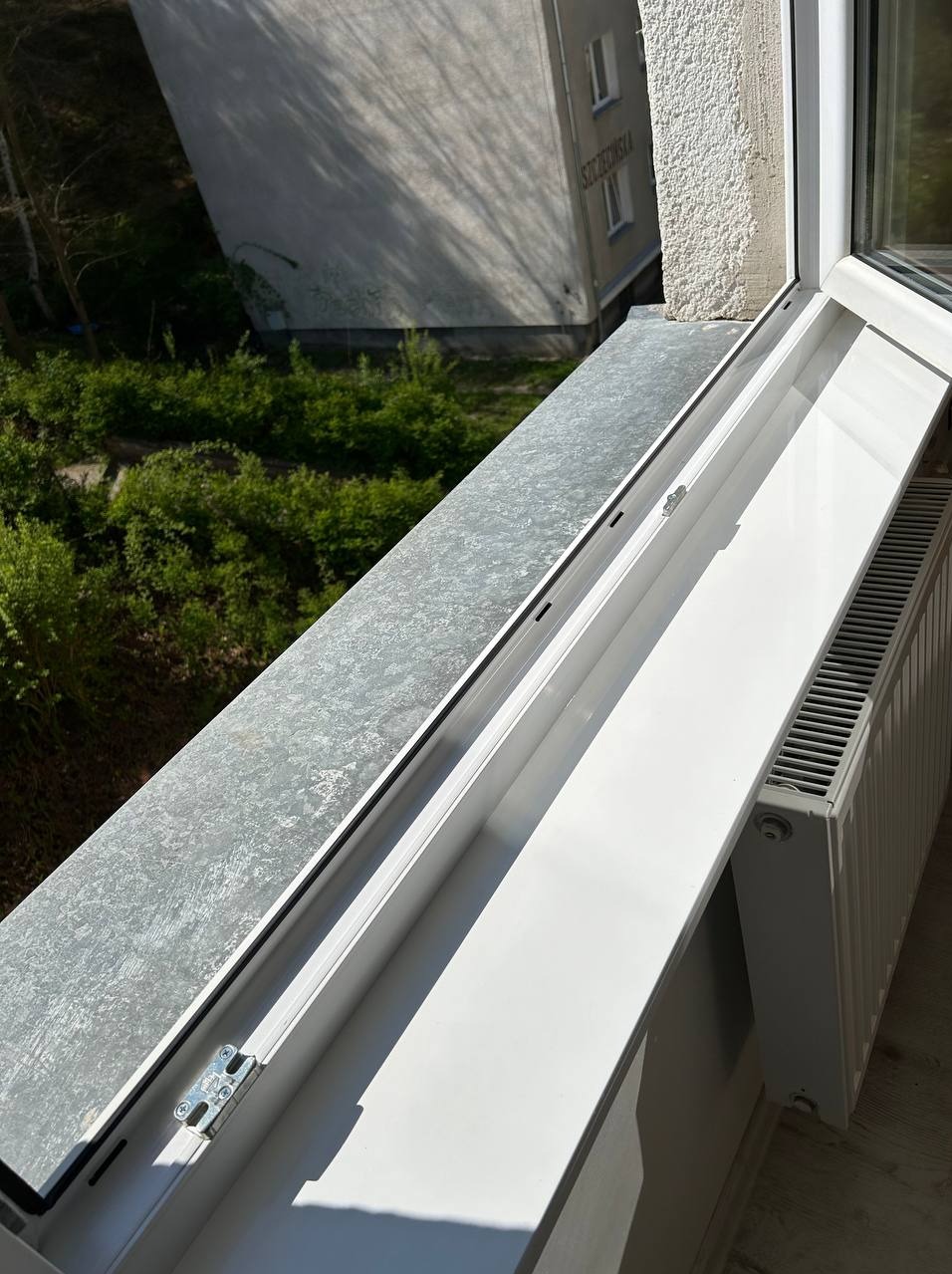 Wnętrze okna PCV z widocznym parapetem zewnętrznym z blachy ocynkowanej, uszczelką, ramą okienną i grzejnikiem, z widokiem na zieleń i budynek mieszkalny w tle.