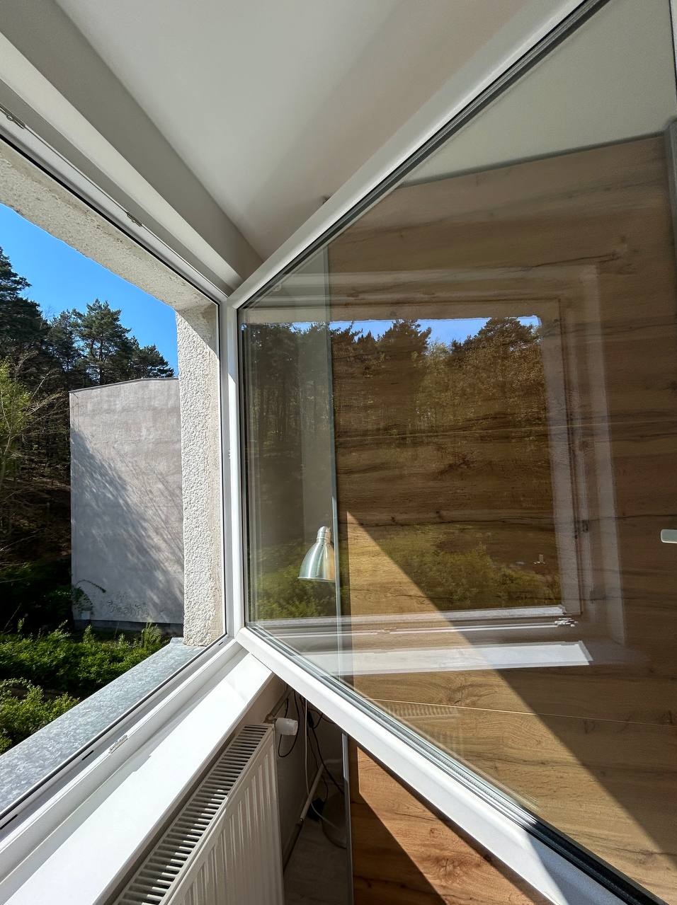 Otwarty narożny segment okna z widokiem na drzewa i budynek w tle, widoczny grzejnik pod parapetem, odbicie lampy na szybie.