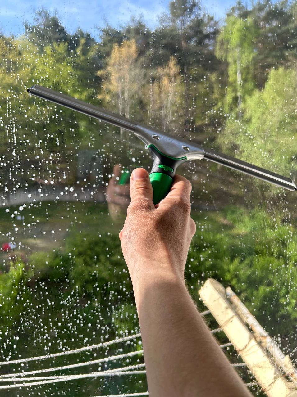 Ręka z zielonym uchwytem ściągaczki do okien na tle szyby pokrytej pianą, z widokiem na zieleń drzew i fragment drabiny sznurkowej w tle.