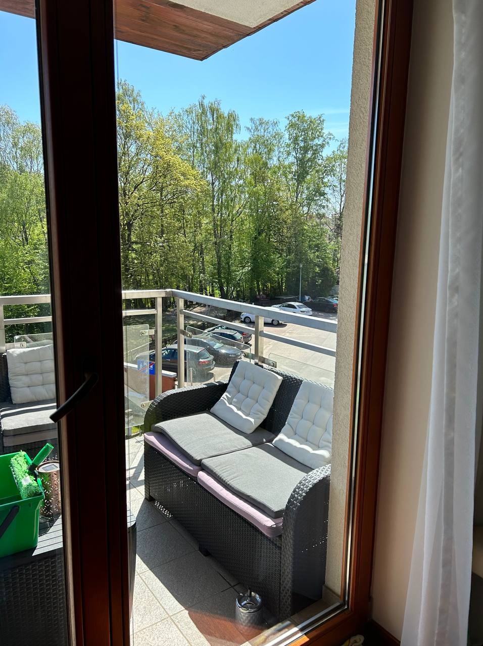 Widok z balkonu na zieleń drzew, meble ogrodowe z szarymi poduszkami i białą zasłoną w oknie, w tle ulica z zaparkowanymi autami.