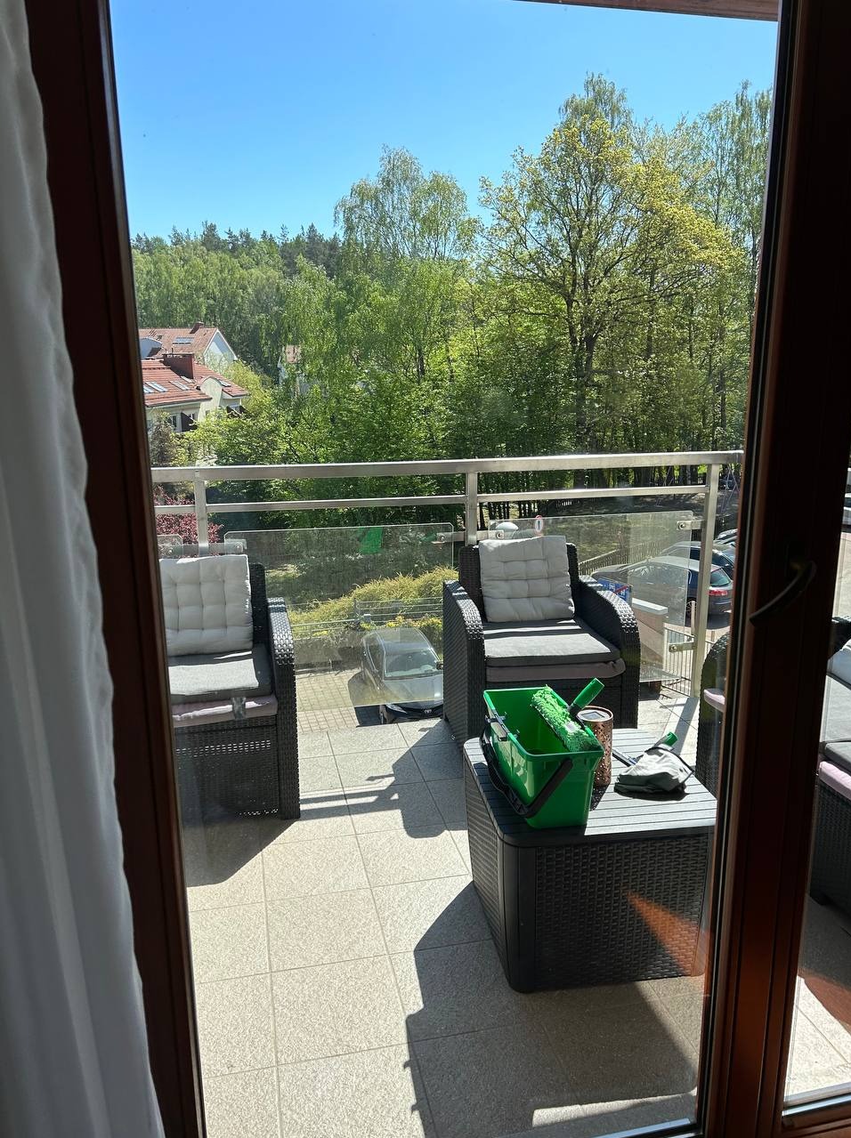 Zestaw do mycia okien w zielonym wiadrze na stoliku balkonowym, widok na zieleń drzew i zabudowę przez szklaną balustradę, słoneczny dzień.