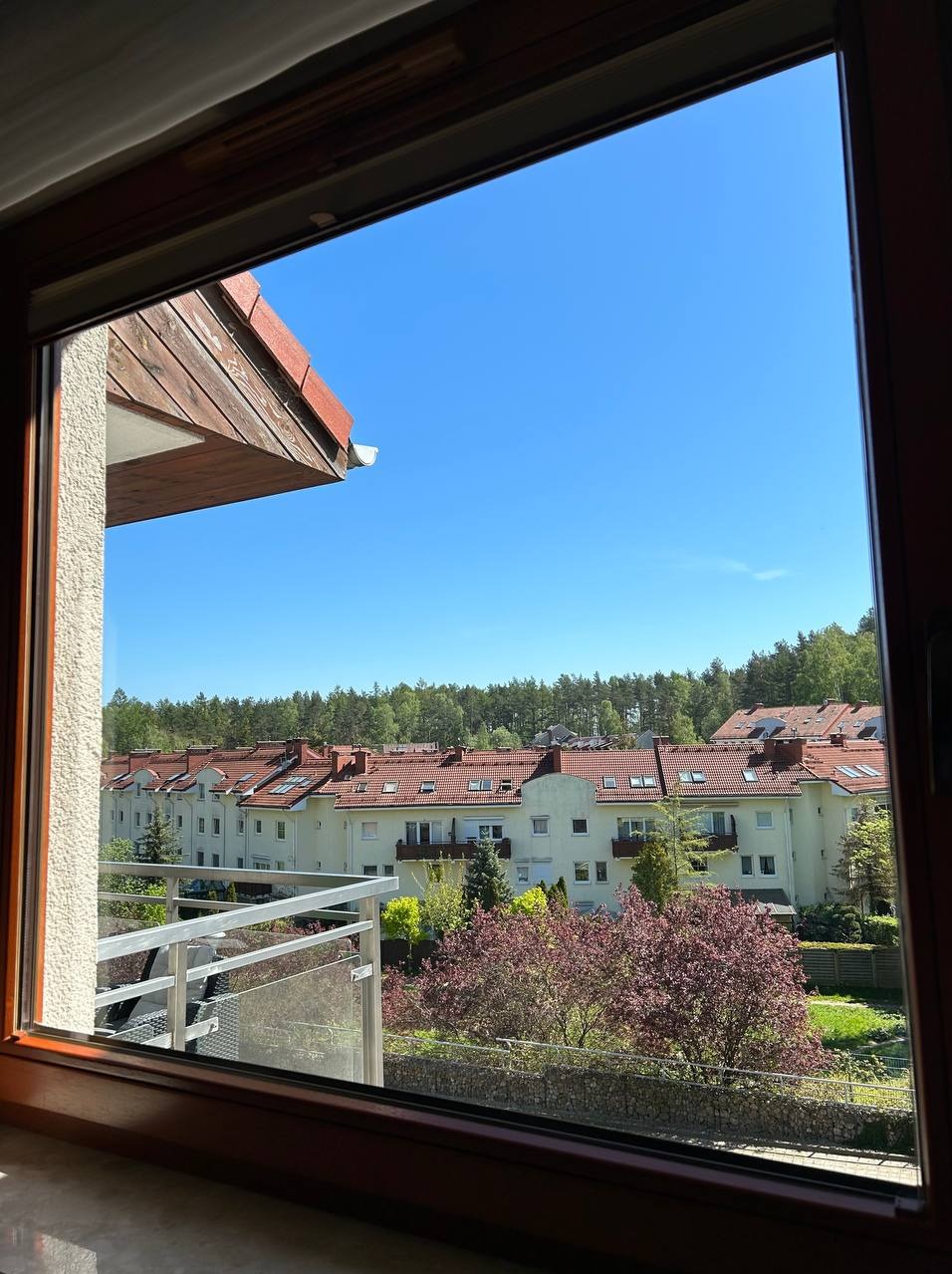 Widok z okna na osiedle domów z czerwonymi dachami w słoneczny dzień. Na pierwszym planie rama okienna z drewna.