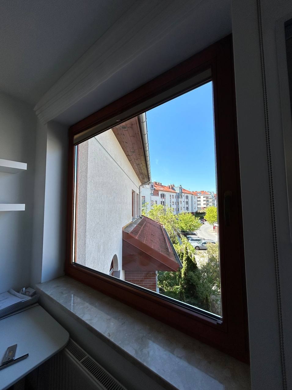 Widok z okna na budynek z czerwoną dachówką i zieleń drzew w słoneczny dzień, rama okienna z ciemnego drewna, marmurowy parapet.