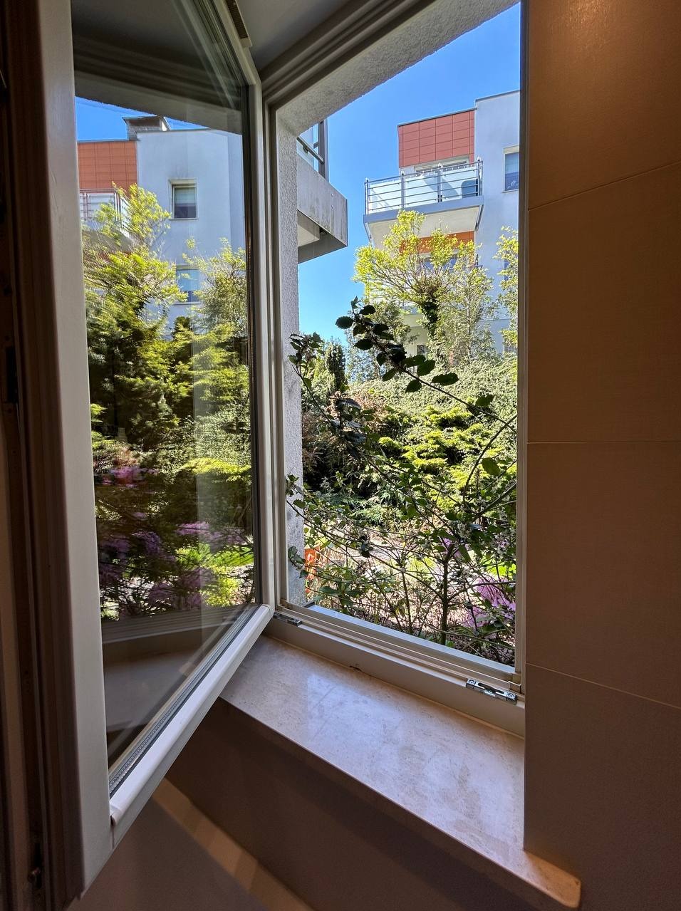 Otwarty, uchylony fragment okna z widokiem na zieleń, budynki w oddali i błękitne niebo; parapet z marmurowym wzorem.