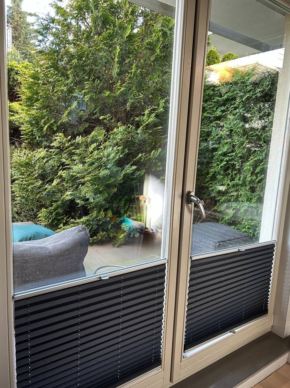 Zasłonięte plisami okno tarasowe z widokiem na zielony ogród z elementami drewnianego tarasu i mebli ogrodowych