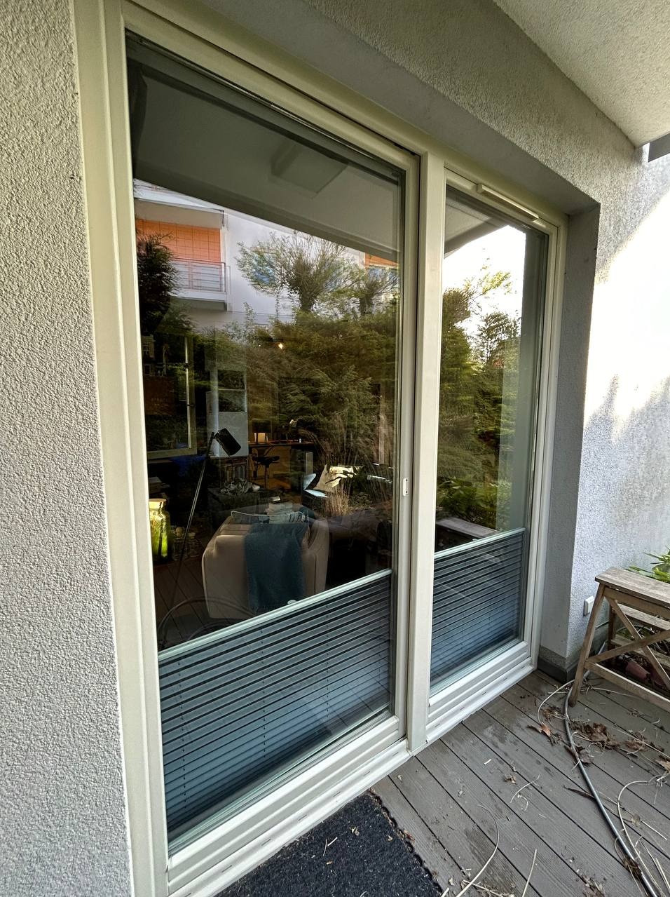 Przesuwne okno balkonowe z widocznymi roletami plisowanymi, na drewnianym balkonie z resztkami liści, odbijające drzewa i budynek z pomarańczowym balkonem.