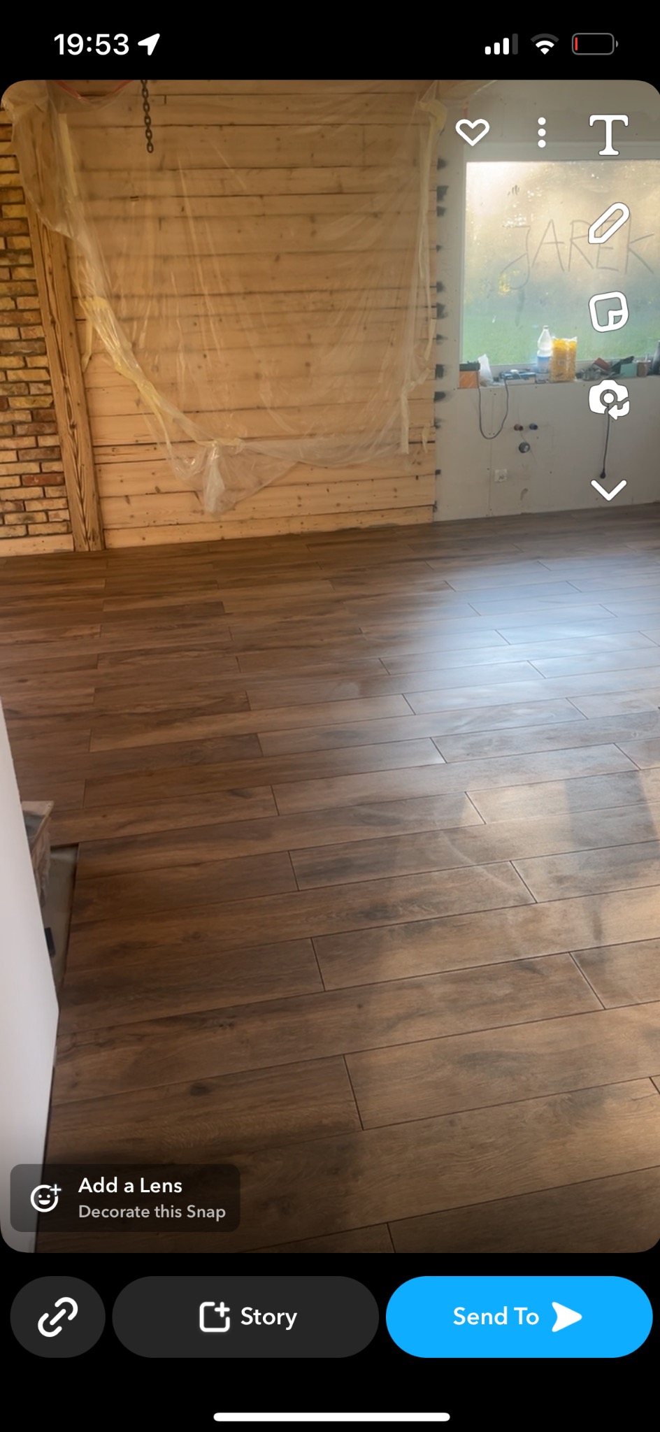 Ułożone brązowe płytki podłogowe imitujące drewno w trakcie remontu, ściana z drewnianych paneli częściowo zabezpieczona folią malarską, okno z napisem 'JAREK'.