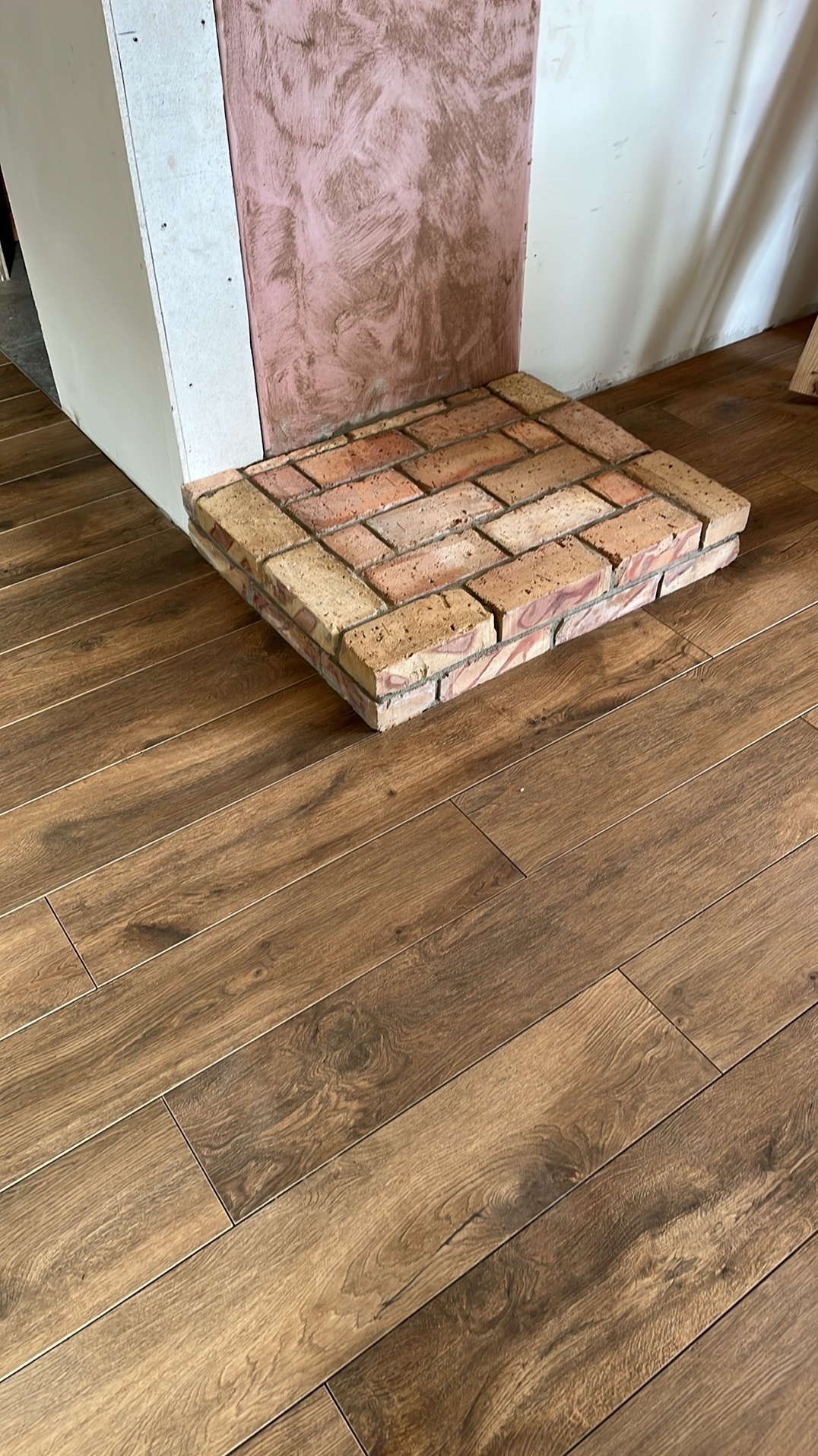 Wykończenie kominka z cegły klinkierowej na podłodze z paneli imitujących drewno, widoczna ściana w trakcie prac wykończeniowych.