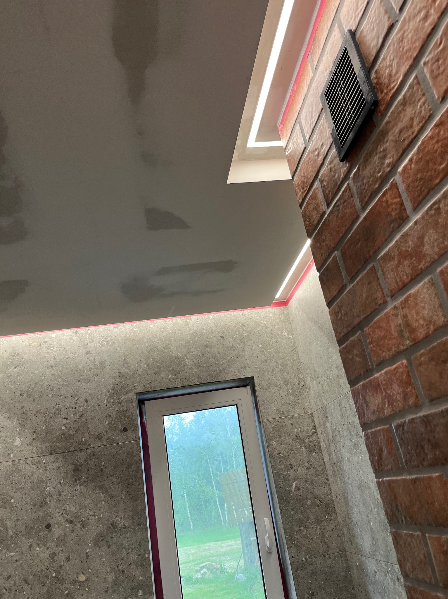 Wykończona łazienka z szarymi płytkami imitującymi kamień, ceglaną ścianą z wentylatorem, oświetleniem LED w suficie podwieszanym i oknem z widokiem na zieleń.