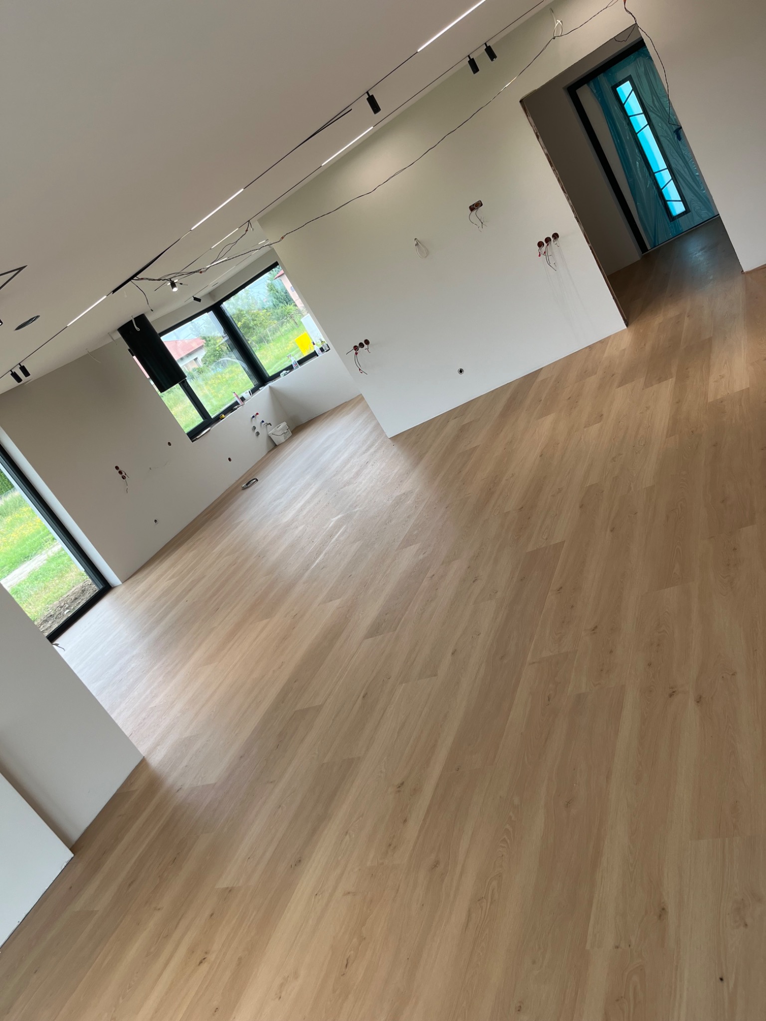Jasne panele podłogowe ułożone w pustym pokoju z białymi ścianami, widoczne okna z czarnymi ramami, instalacja elektryczna w trakcie montażu, drzwi wejściowe zabezpieczone folią.