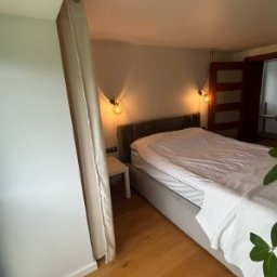 Fhu Ren Dam - Sypialnia w Krakowie po remoncie: drewniana podłoga, szara tapicerowana rama łóżka, lampki ścienne i zasłona oddzielająca przestrzeń. Jasne, ciepłe oświetlenie.