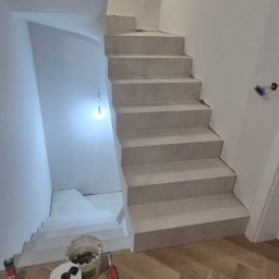 Fhu Ren Dam - Wykończone schody w jasnej tonacji, z widocznymi narzędziami i materiałami budowlanymi na parkiecie u dołu. Minimalistyczne wnętrze w trakcie prac wykończeniowych.