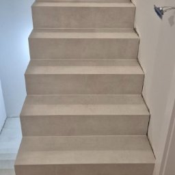 Fhu Ren Dam - Schody wykończone jasnoszarym gresem, widok z dołu na drewnianą podłogę ułożoną w jodełkę. Ściana biała, widoczne niedociągnięcia przy wykończeniu.