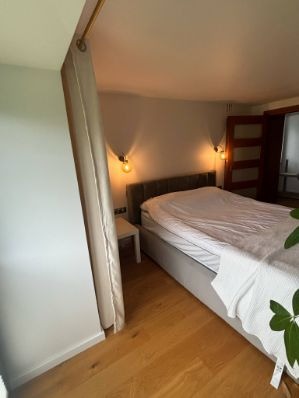 Sypialnia w Krakowie po remoncie: drewniana podłoga, szara tapicerowana rama łóżka, lampki ścienne i zasłona oddzielająca przestrzeń. Jasne, ciepłe oświetlenie.