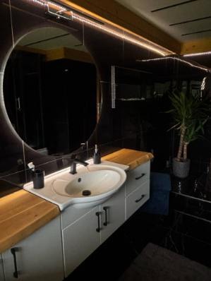 Nowoczesna łazienka w czerni z drewnianym blatem i okrągłym lustrem podświetlonym LED. Biała szafka pod umywalką z czarnymi uchwytami. Roślina doniczkowa w tle.