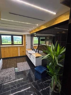 Nowoczesna łazienka z czarną marmurową podłogą, drewnianymi panelami ściennymi i oknem z widokiem na zieleń. Umywalka z owalnym lustrem i oświetleniem LED.