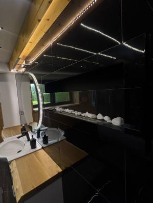 Nowoczesna łazienka z czarnymi płytkami, drewnianym blatem i podświetlanym lustrem. Na półce dekoracyjne białe kamienie. Oświetlenie LED na suficie.