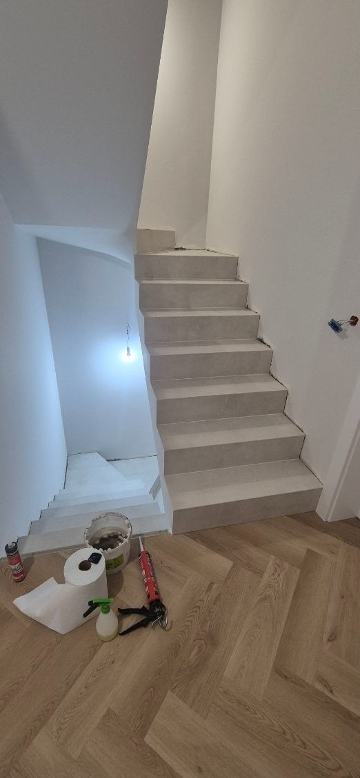 Wykończone schody w jasnej tonacji, z widocznymi narzędziami i materiałami budowlanymi na parkiecie u dołu. Minimalistyczne wnętrze w trakcie prac wykończeniowych.
