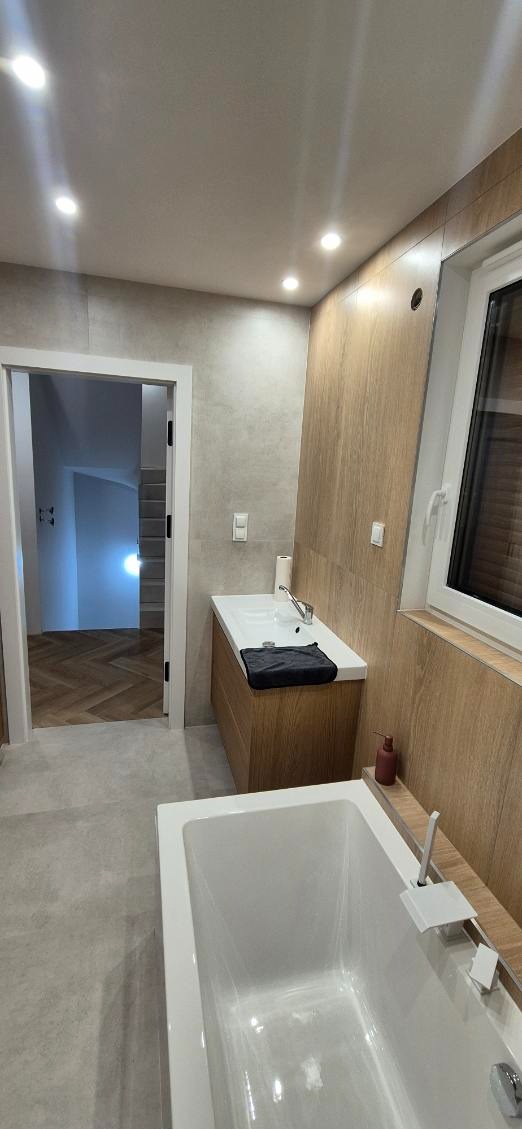 Nowoczesna łazienka po remoncie: biała wanna, umywalka z drewnianą szafką, szare płytki i drewnopodobne panele ścienne. Widok na korytarz z parkietem.