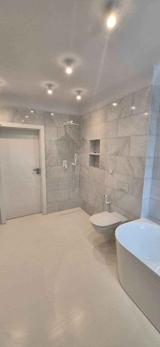 Jasna łazienka po remoncie: płytki imitujące kamień, biała armatura (wanna, toaleta), podwieszana deszczownica i oświetlenie punktowe. Minimalistyczny design.