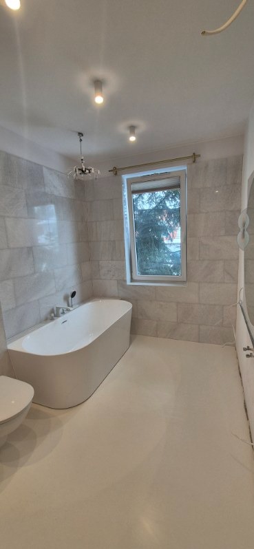 Nowoczesna łazienka z wolnostojącą wanną, marmurowymi płytkami na ścianach i jasną podłogą żywiczną. Okno z widokiem na zieleń. Minimalistyczny design i oświetlenie.