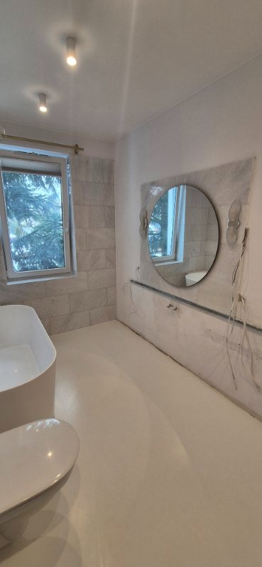 Jasna łazienka po remoncie: biała wanna, toaleta, okrągłe lustro z dekoracjami, płytki imitujące kamień na ścianie, gładka podłoga. Widoczne niedokończone podłączenia.